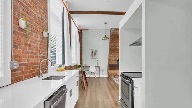 Una cocina con pared de ladrillo, muebles blancos, un lavamanos de acero inoxidable y un horno empotrado.