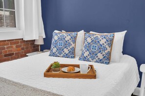 Una cama bien hecha con edredón blanco, dos almohadas azules estampadas, una bandeja de madera con un croissant, una taza y una pequeña planta.