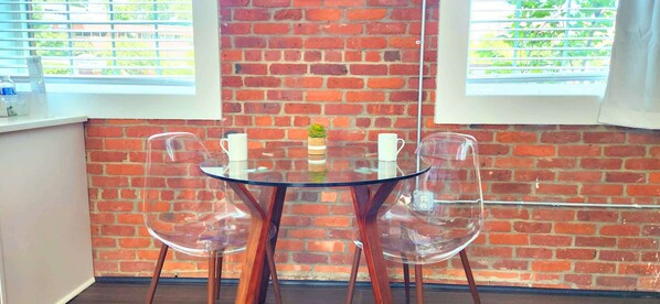 Une table en verre, entourée de quatre chaises, se trouve dans une pièce dont les murs sont en briques et qui possède des fenêtres.