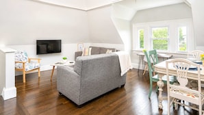 Een moderne woonkamer met een grijze bank, houten stoelen, een eettafel en een aan de muur bevestigde televisie.