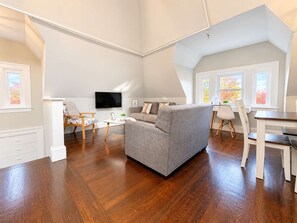 A spacious living area with a grey sofa, wooden flooring, a flat-screen TV, and a dining table with chairs. - Jennifer’s Unique Loft Near Brown 2 Parking Spots (Providence)