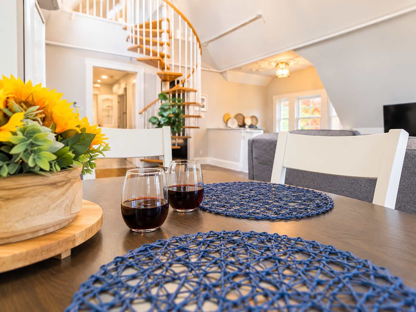 A dining area with a wooden table, two wine glasses, a blue woven placemat, and a sunflower arrangement.