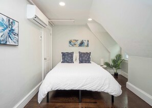 Een slaapkamer met een wit bed, twee gestreepte kussens, een schilderij en een potplant.