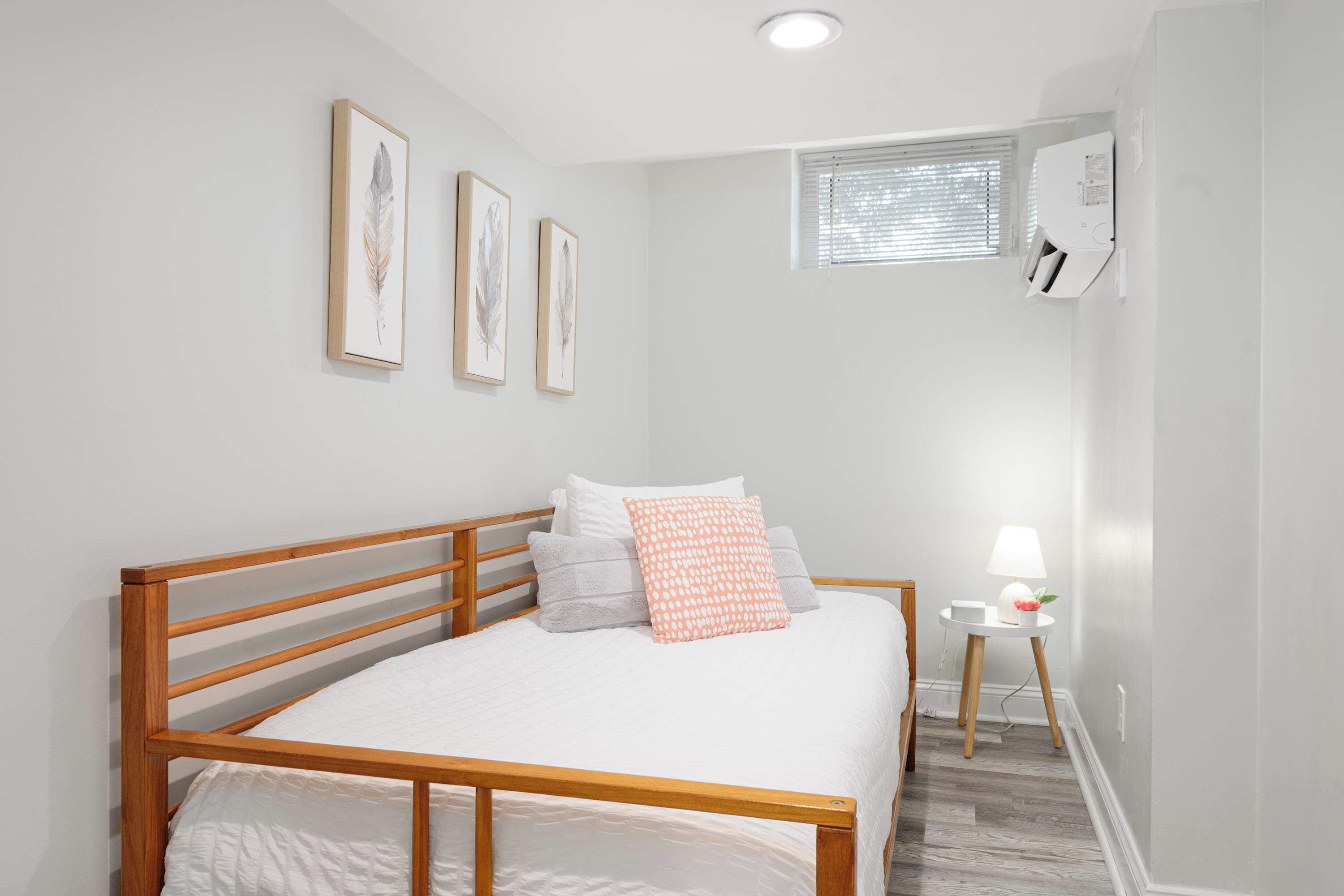 A bedroom with a wooden bed frame, white bedding, a small bedside table with a lamp, and framed artwork on the wall.
