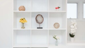 A white shelving unit with decorative items including a woven basket, a flower arrangement, a round object, and a potted plant.