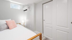 A bedroom with a bed, a nightstand, a lamp, a window with blinds, an air conditioning unit, and a door. - Jennifer’s Walk to Brown U. Private Entry & Patio (Providence)