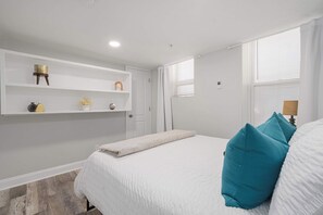 Een slaapkamer met een bed, een witte muur, een plank met decoratieve objecten en een raam met zonwering.