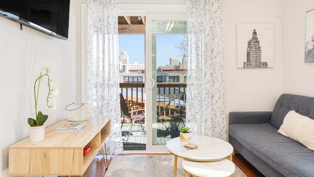 A living room with a sofa, a small round table, a TV mounted on the wall, and a view through a glass door.