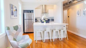 A modern kitchen with a central island, white bar stools, and a stainless steel refrigerator.