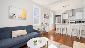 A modern living room with a sofa, a small round table with a plant, and a kitchen area with white cabinets and bar stools.