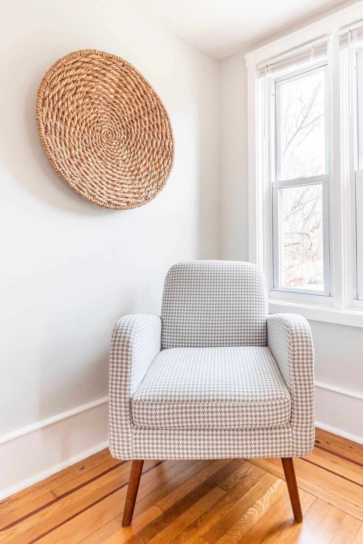 A chair with a houndstooth pattern, a woven wall hanging, and a window with a view of bare trees.