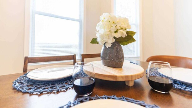 Een eettafel met een vaas witte bloemen, twee glazen rode wijn en een geweven placemat.