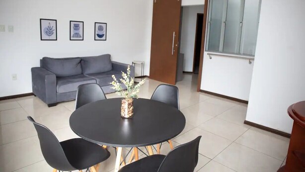 A modern living room with a grey sofa, a round black table with four chairs, and a vase with flowers. There are three framed pictures on the wall.