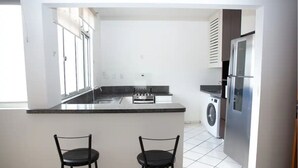 A compact kitchen with a central island, two bar stools, a washing machine, and a refrigerator.