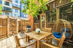 Une terrasse extérieure avec une table et des chaises en bois, un fauteuil suspendu en osier et un mur en briques.