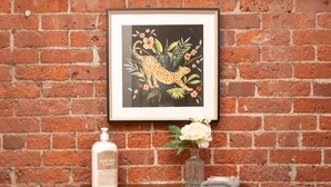 A framed artwork of a leopard with flowers on a brick wall, a bottle of goat milk, and a vase with flowers on a wooden shelf.