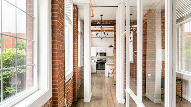 A narrow hallway with brick walls, large windows, and a kitchen area in the background.