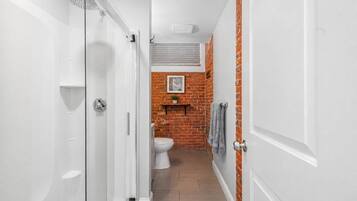 A narrow hallway leading to a bathroom with a shower, toilet, and a brick wall with a shelf and a picture.