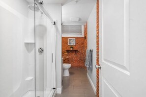 Un pasillo estrecho que conduce a un baño con ducha, inodoro y una pared de ladrillo con una estantería y un cuadro.