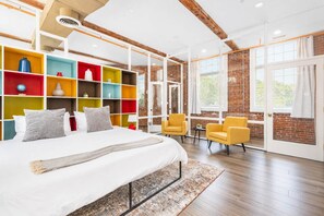 A modern bedroom with a large bed, colorful built-in shelving, a wooden floor, and a brick wall. - Jennifer’s Historic Loft | Glass-Enclosed Bedroom (Providence)