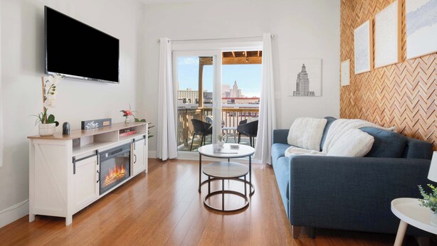 A modern living room with a fireplace, a flat-screen TV, a sofa, and a balcony with city views. - Jennifer’s Chic Apt w/ Downtown City Views Private Deck (Providence)