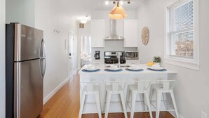 A modern kitchen with a stainless steel refrigerator, white bar stools, a central island with a sink, and a pendant light. - Jennifer’s Chic Apt w/ Downtown City Views Private Deck (Providence)