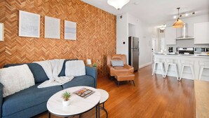 A modern living room with a herringbone wood wall, a sofa, a coffee table, a reading chair, and a kitchen area with white bar stools. - Jennifer’s Chic Apt w/ Downtown City Views Private Deck (Providence)