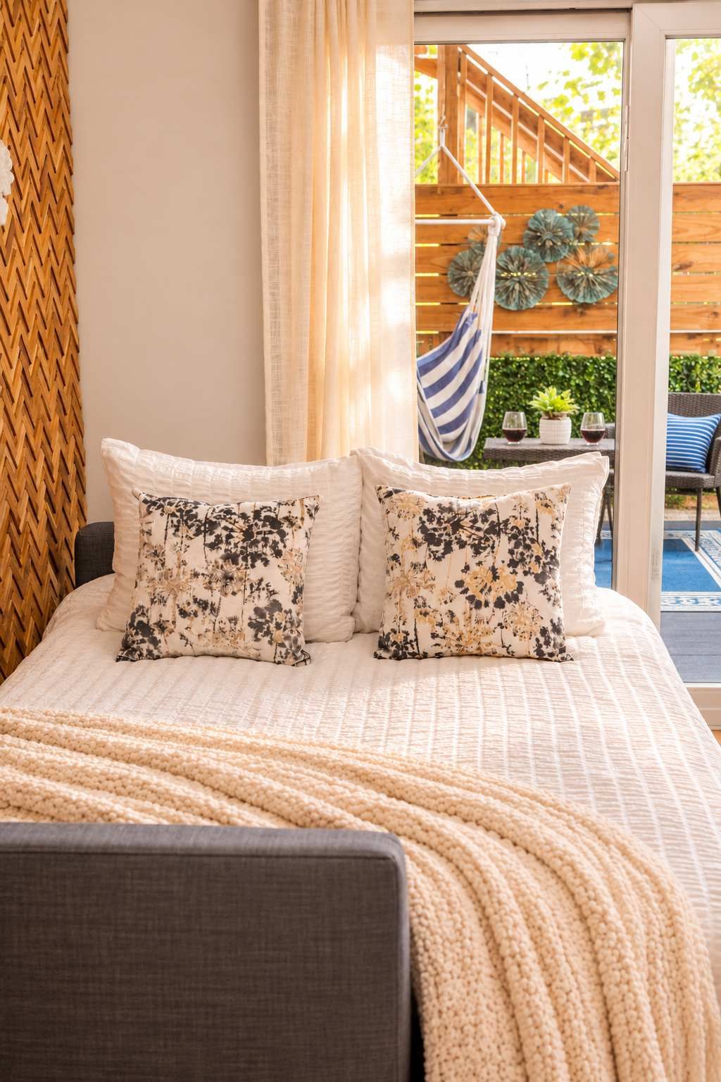 A bed with floral patterned pillows, a knitted throw blanket, and a view of an outdoor wooden deck and plants.