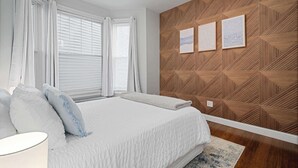 Ein Schlafzimmer mit einem Bett, weißen Vorhängen, einer Holzwand mit geometrischen Mustern und drei gerahmten Bildern.