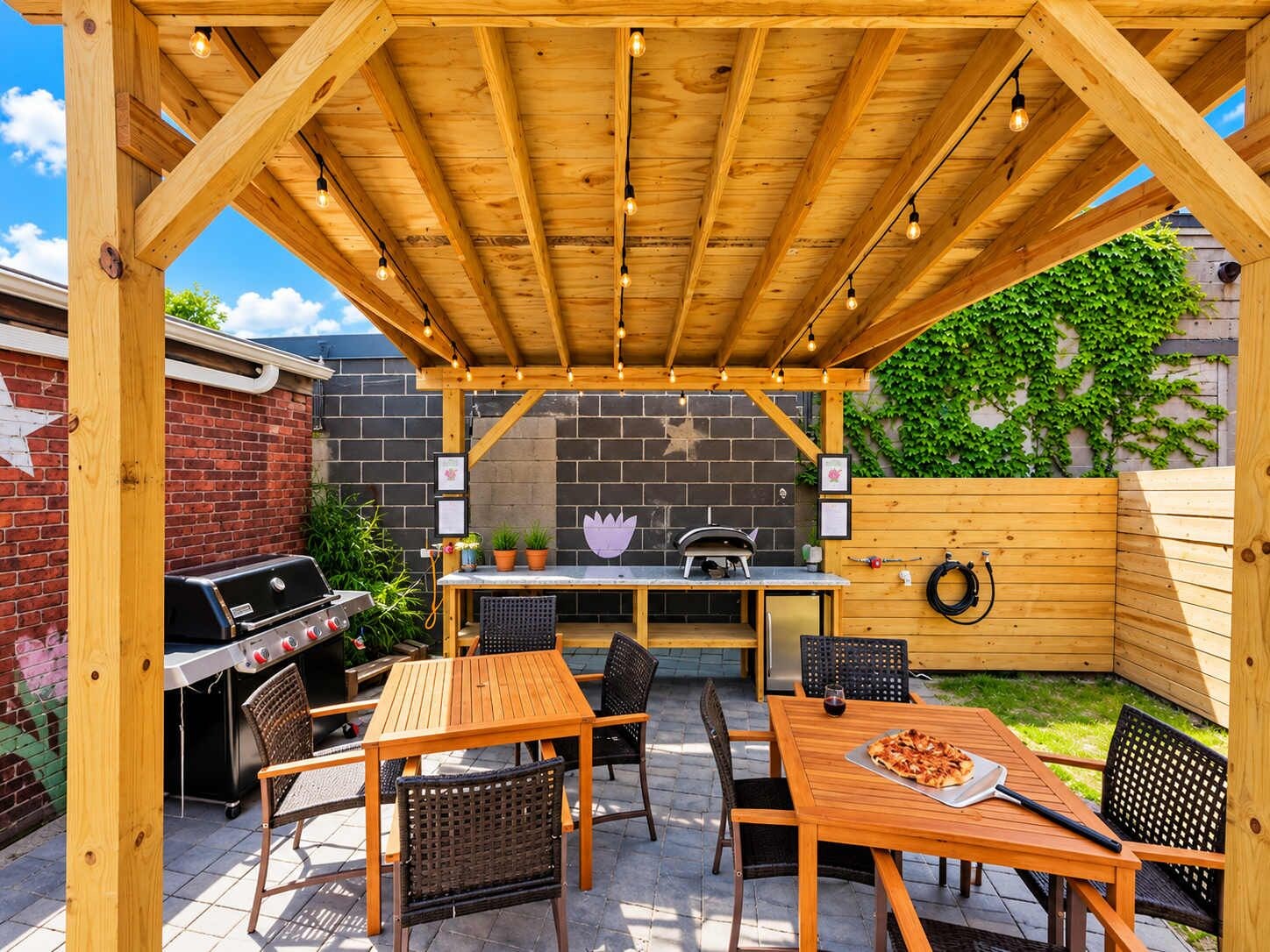 An outdoor patio with a wooden pergola, a barbecue grill, a table with chairs, and a pizza on the table.