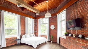 A room with a bed, large windows, a TV mounted on the wall, and a clock. - Jennifer’s Historic Penthouse Loft | City Views (Providence)