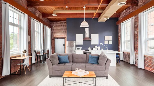 A spacious living area with a gray sofa, wooden ceiling beams, and large windows. - Jennifer’s Historic Penthouse Loft | City Views (Providence)