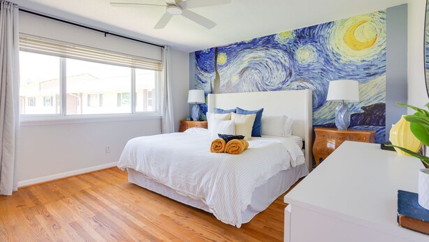 Une chambre à coucher avec un grand lit, deux tables de chevet, un ventilateur de plafond et une fenêtre avec des stores.