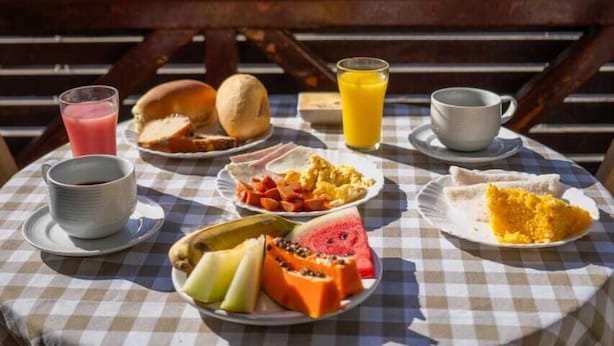 A table with breakfast items including bread, fruit, and a drink, set outdoors with a bench and trees in the background. - Pe na Areia Pousada Jeri (Jijoca de Jericoacoara)