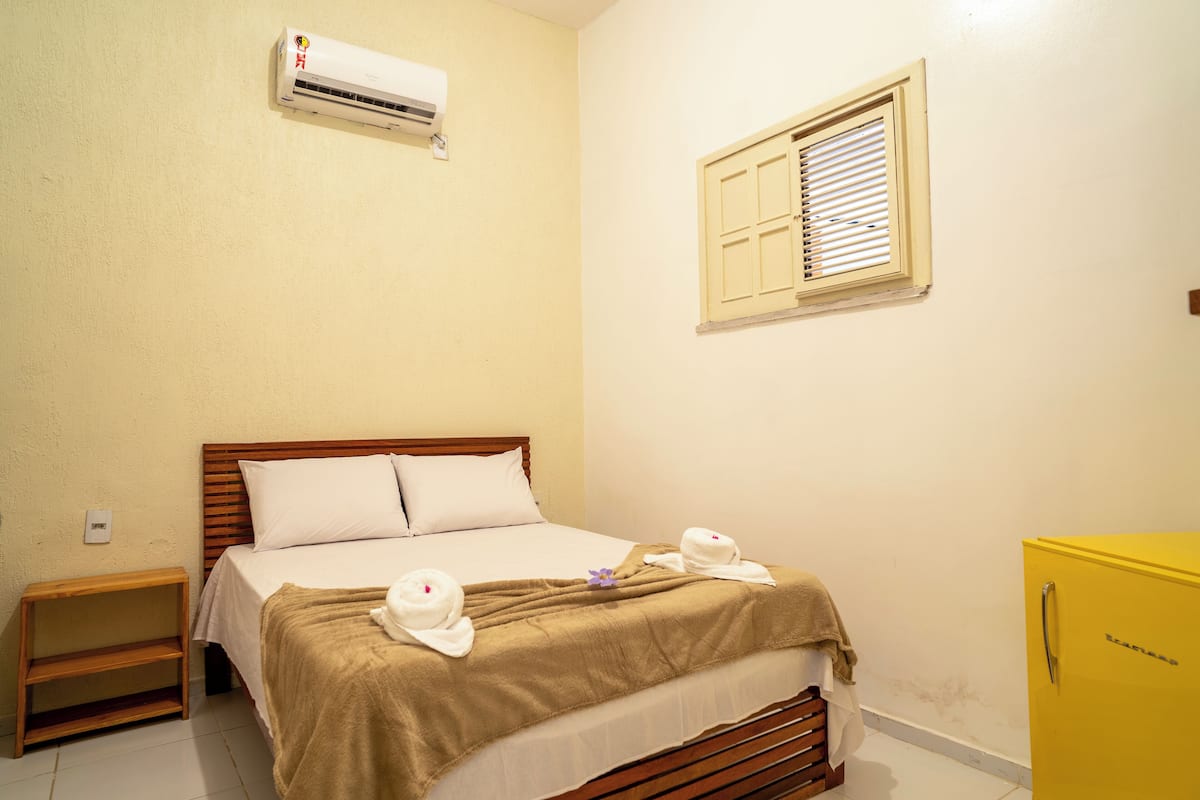 Une chambre d’hôtel avec un lit, une table de chevet, un climatiseur, une fenêtre avec des volets et une armoire jaune.