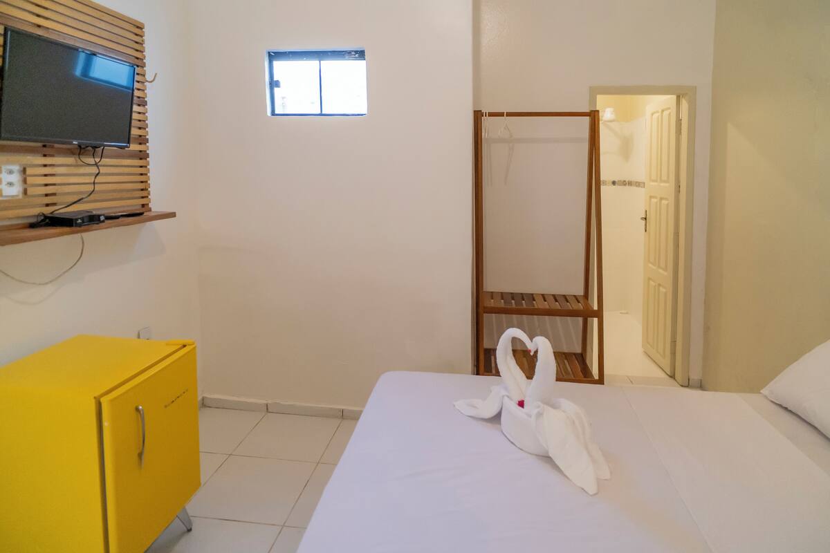 Une chambre d’hôtel avec un lit, un téléviseur posé sur une étagère en bois, une armoire jaune et une fenêtre.