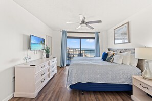 Un dormitorio con cama, cómoda, ventilador de techo, televisor, balcón con vistas y un cuadro en la pared.
