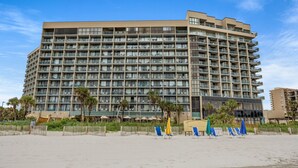Een hotel met meerdere verdiepingen en balkons, gelegen nabij een zandstrand met ligbedden en parasols.