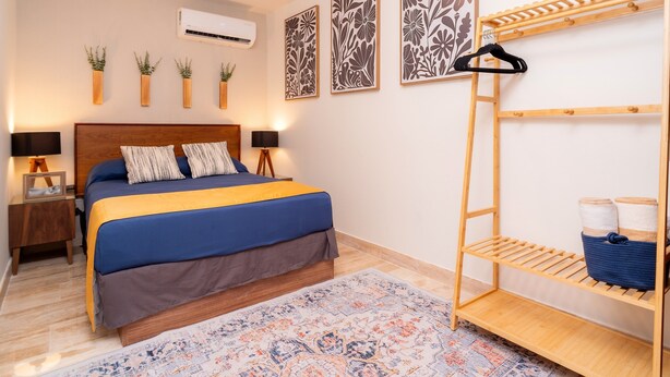 A bedroom with a bed, bedside tables, a wooden shelf unit, and decorative wall art. - AWA Playacar 2BR - Resort Pools, Balcony, Walk to Beach (Playa del Carmen)
