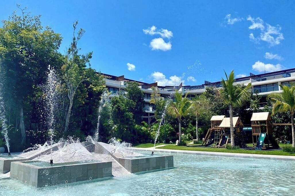 Une piscine agrémentée de jeux d’eau, entourée de palmiers et bordée d’un bâtiment doté de nombreux balcons.