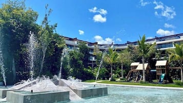 Une piscine agrémentée de jeux d’eau, entourée de palmiers et bordée d’un bâtiment doté de nombreux balcons.