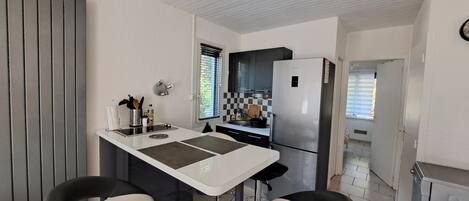 Cocina compacta con una isla central, dos taburetes de bar, un refrigerador y una ventana con persianas.