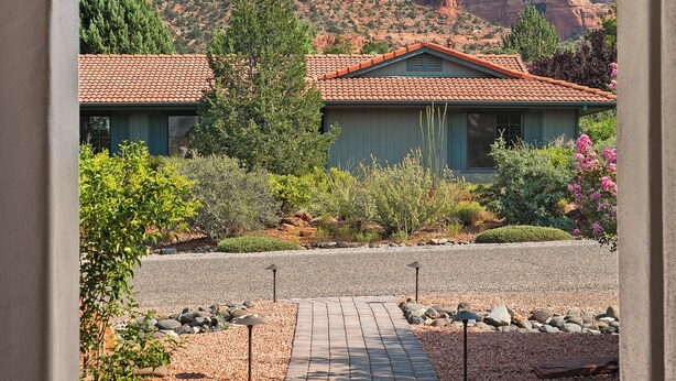 A view through an arched doorway of a building with a red-tiled roof, surrounded by a landscaped garden and a paved pathway leading to it.