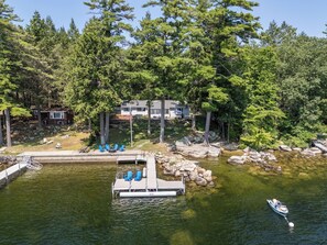 A lakeside property with a dock, outdoor seating, and a house surrounded by trees. - Direct Sebago Lakefront – Beach, Dock, Mooring, 3BR/2BA + EV Charger (Raymond)