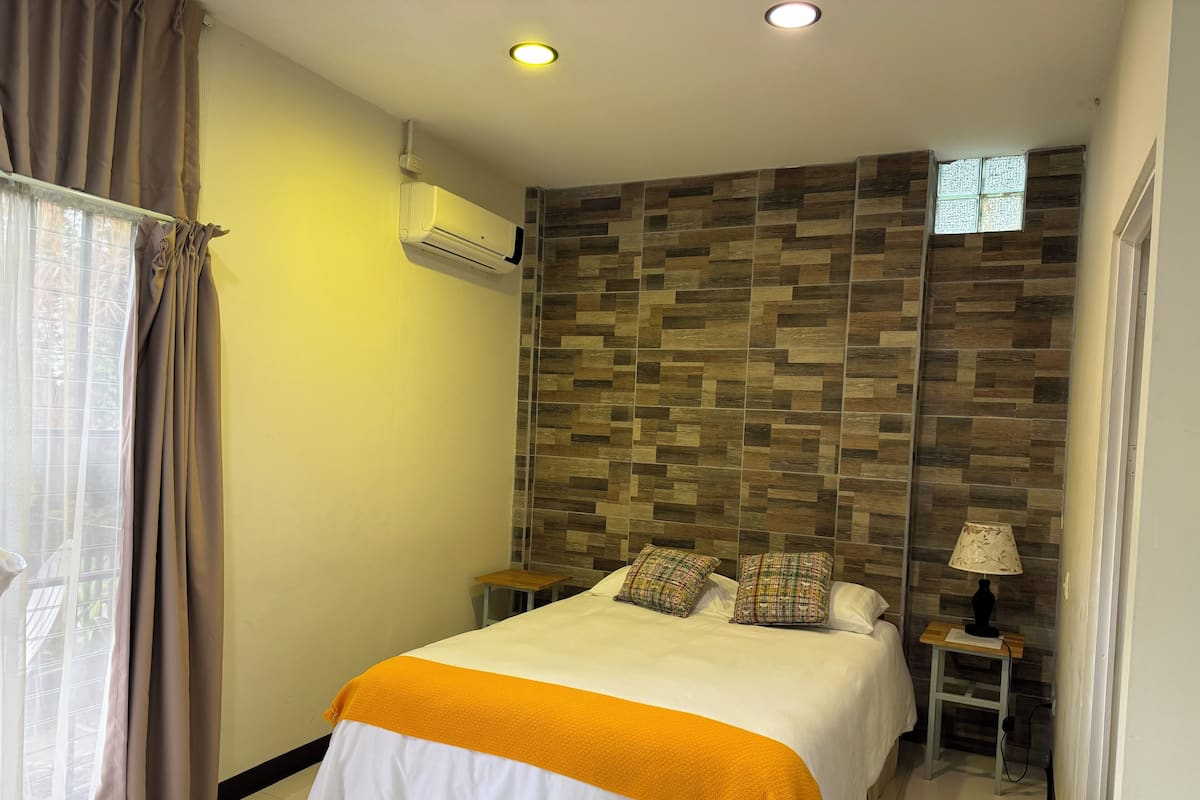 Une chambre à coucher comprenant un lit, une fenêtre avec des rideaux, un appareil de climatisation, une table de chevet, une lampe et un mur orné d’un motif décoratif.