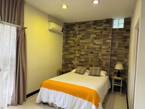 A bedroom with a bed, a window with curtains, an air conditioning unit, a nightstand, a lamp, and a wall with a patterned design. - Legacy Boutique Hotel (Quesada)