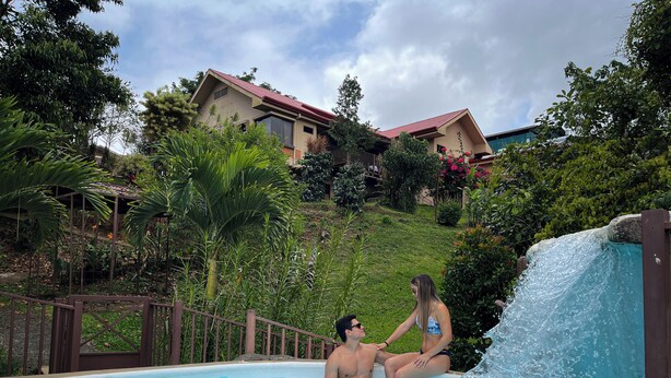 A swimming pool with a waterfall feature, surrounded by lush greenery and a hillside with houses in the background. - Legacy Boutique Hotel (Quesada)