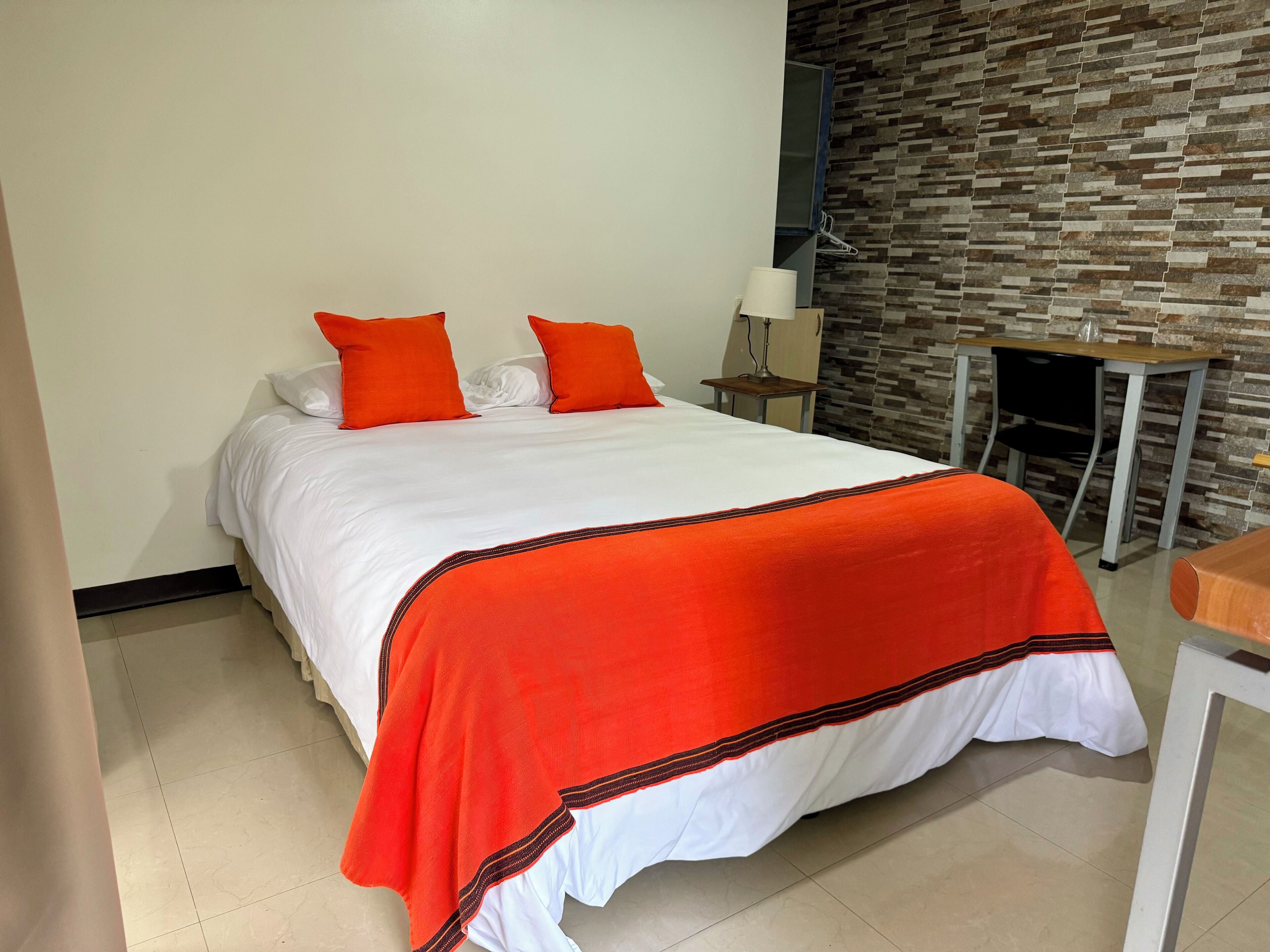 Una cama bien hecha con ropa blanca y almohadas naranjas, una mesita de noche pequeña con una lámpara, y una pared con un patrón de piedra texturizada.