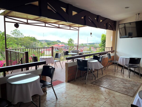 A covered outdoor seating area with tables and chairs, a view of greenery and buildings in the distance, and a tiled floor. - Legacy Boutique Hotel (Quesada)