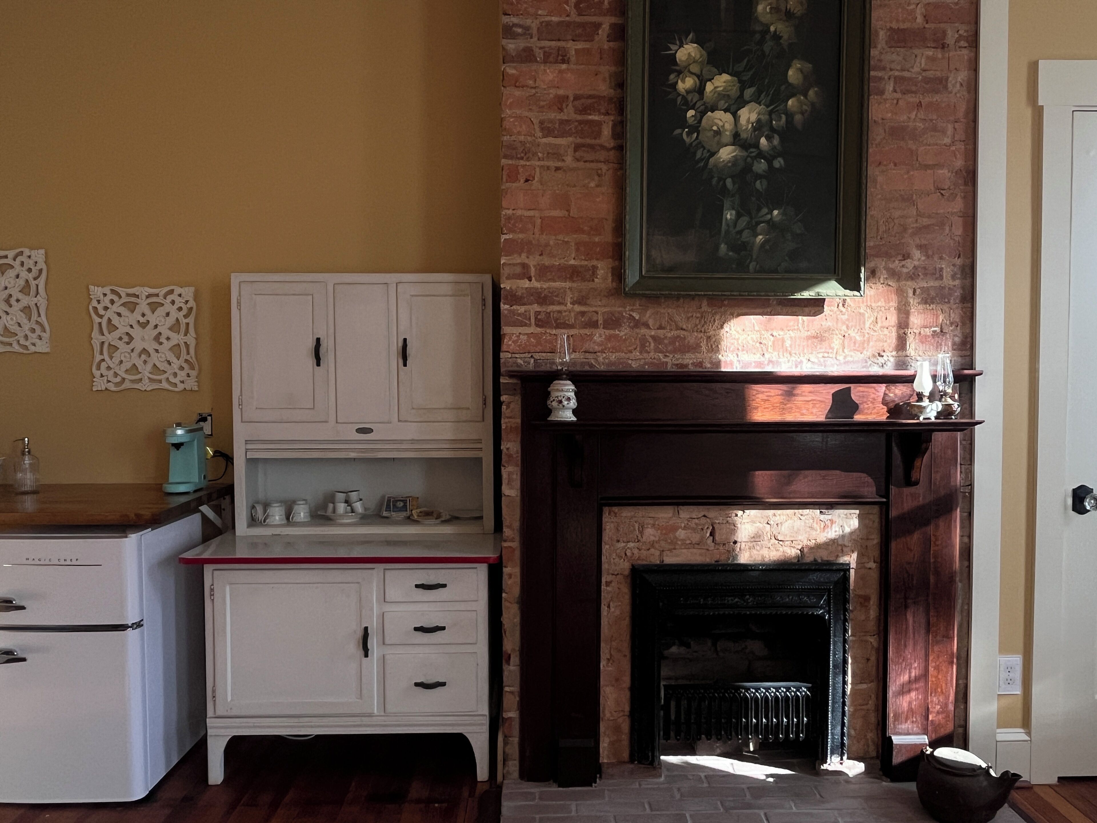 Una cocina con un refrigerador blanco, armarios blancos, una chimenea de ladrillo y un cuadro sobre la chimenea.